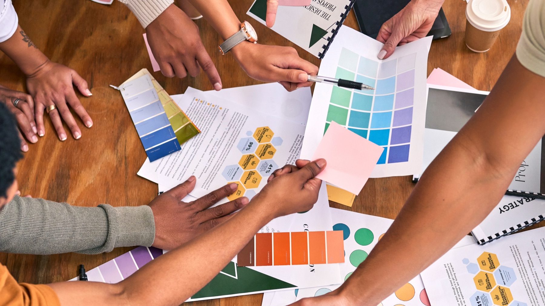 Overhead view of several hands reaching across a table covered in colour swatches, a project proposal booklet, and brand strategy documents