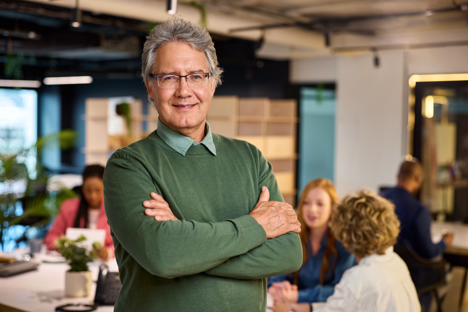 Man standing with arms crossed in an office, with colleagues meeting at a table in the background