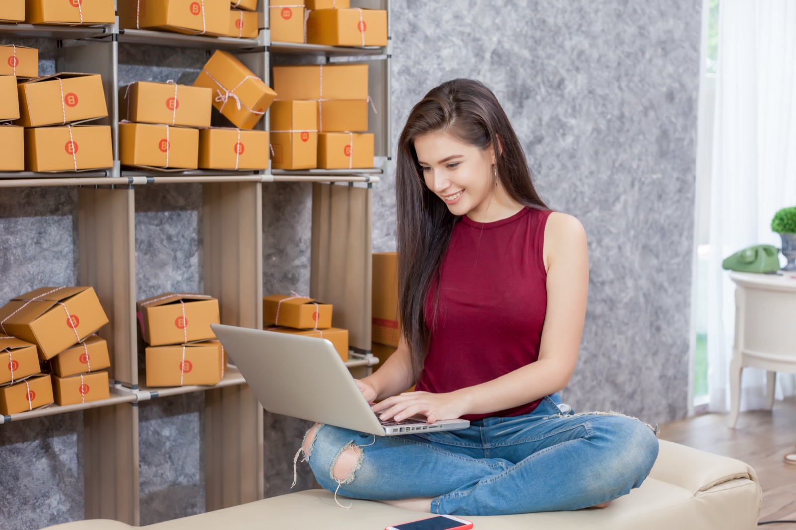 Woman working on a laptop while sitting cross-legged, surrounded by shelves of packed shipping boxes