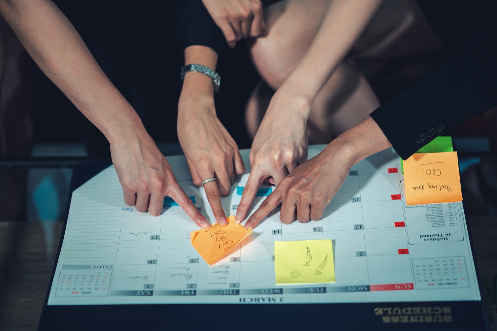 Several hands pointing to a date on a desk calendar covered in sticky notes
